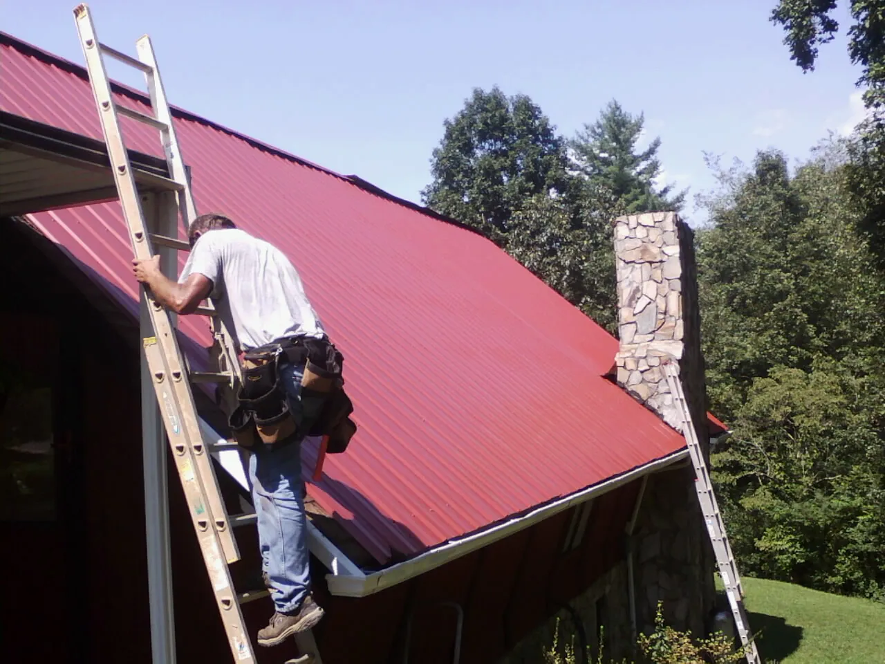 Roofer on a ladder beside a red metal roof and stone chimney — Soffit & Fascia Repair by Roofing Brothers in Red Hill